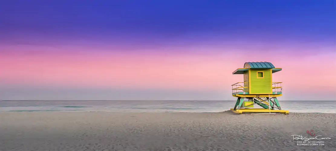 A bright yellow and green lifeguard house sits on the sand of Miami Beach with a pink and purple colored sunset sky