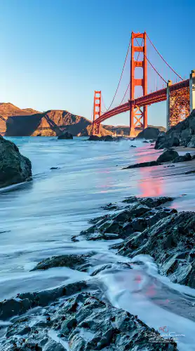 The Golden Gate bridge as seen from the beach with the color reflected on the water.